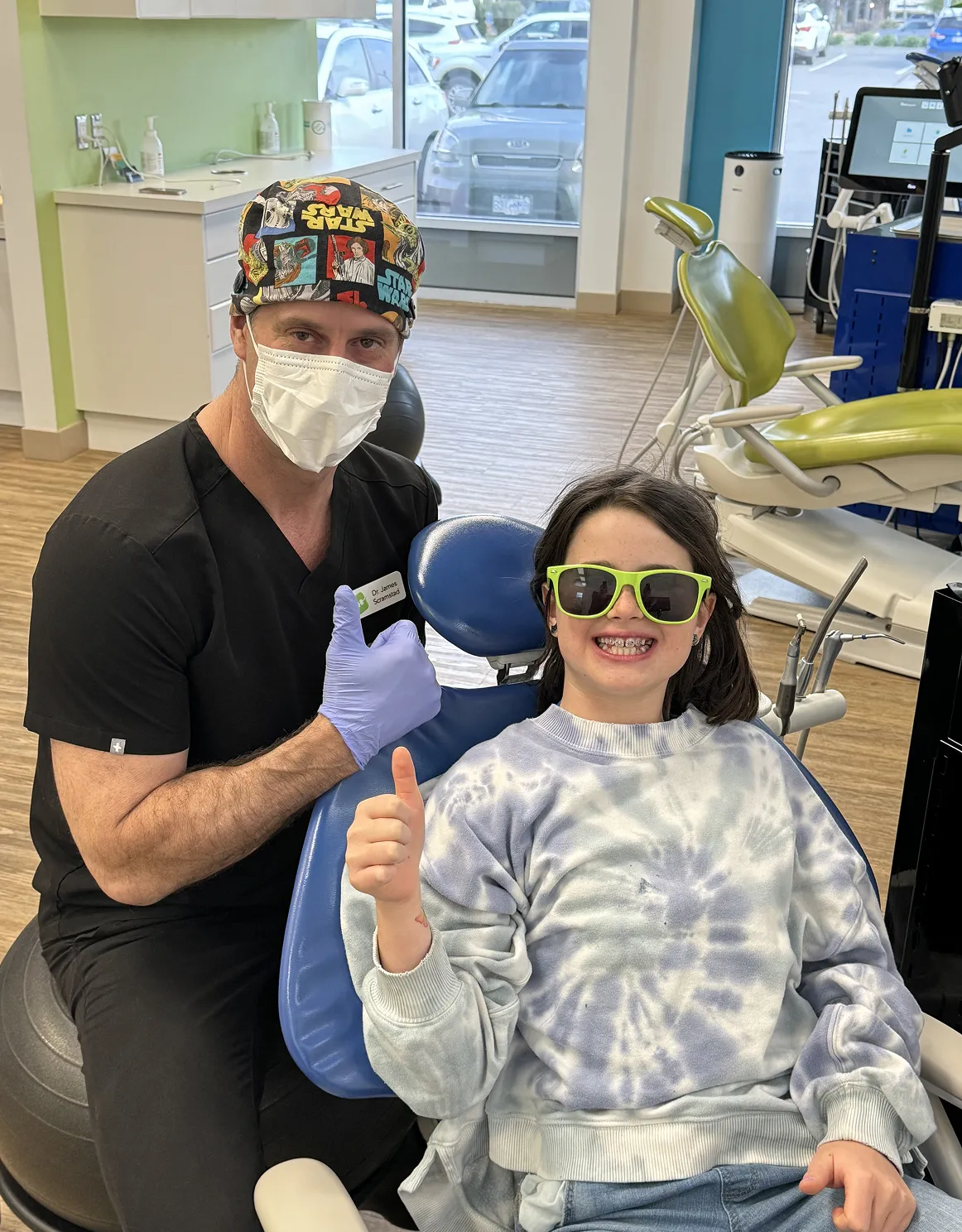 Orthodontist with mask and gloves posing with a young patient wearing sunglasses and braces, both giving thumbs up in a modern dental office, highlighting positive orthodontic experience at Straight Smiles in Kelowna.